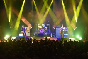A shot from the balcony of The Cribs playing at Tiffcuff Festival at The Roundhouse in London. The three band members are below yellow diagonals of lighting, and the front of the picture shows the silhouettes of the crowd
